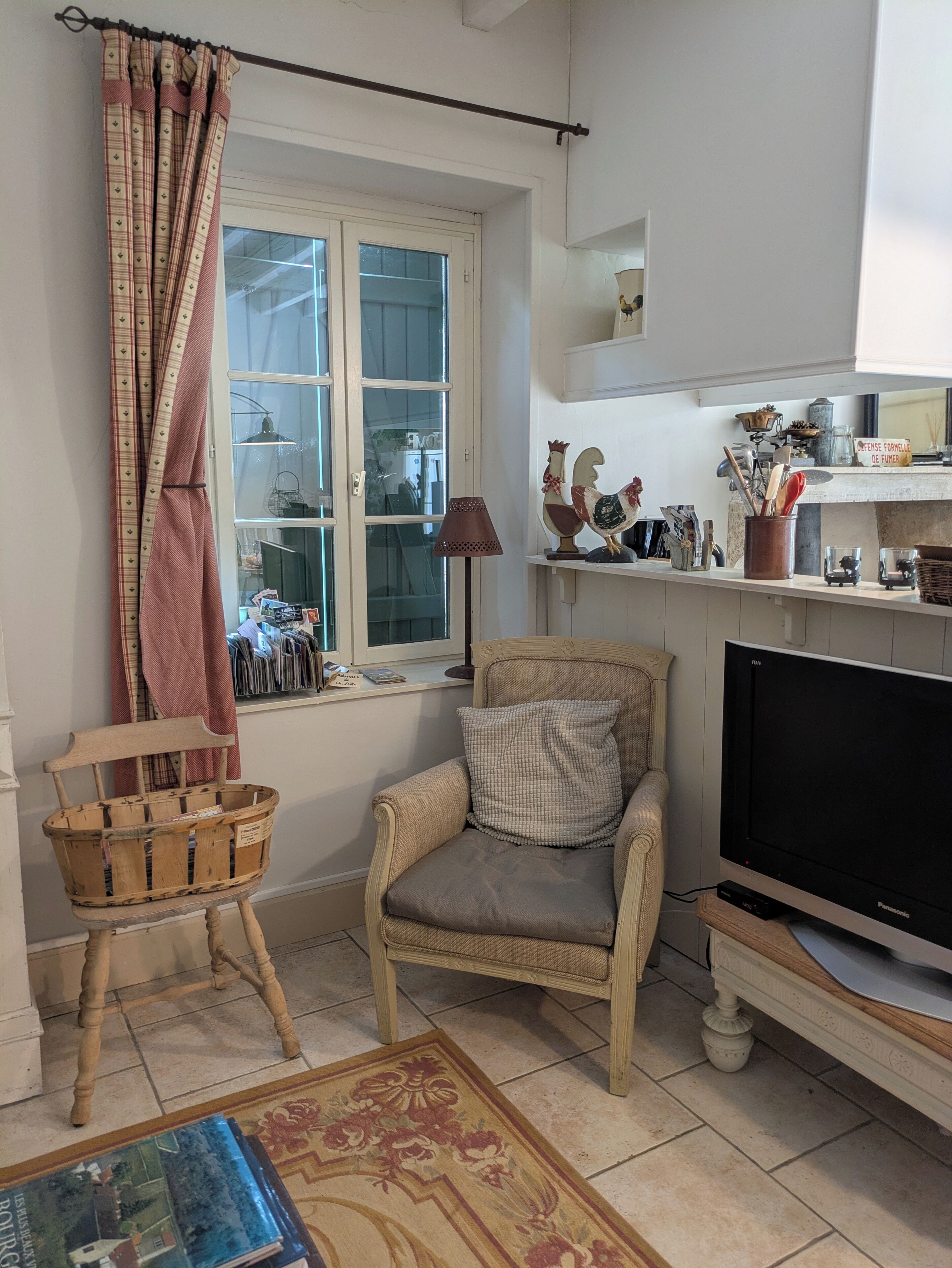 Un salon confortable avec un téléviseur, un fauteuil en bois, une fenêtre avec des rideaux et une étagère contenant divers objets.