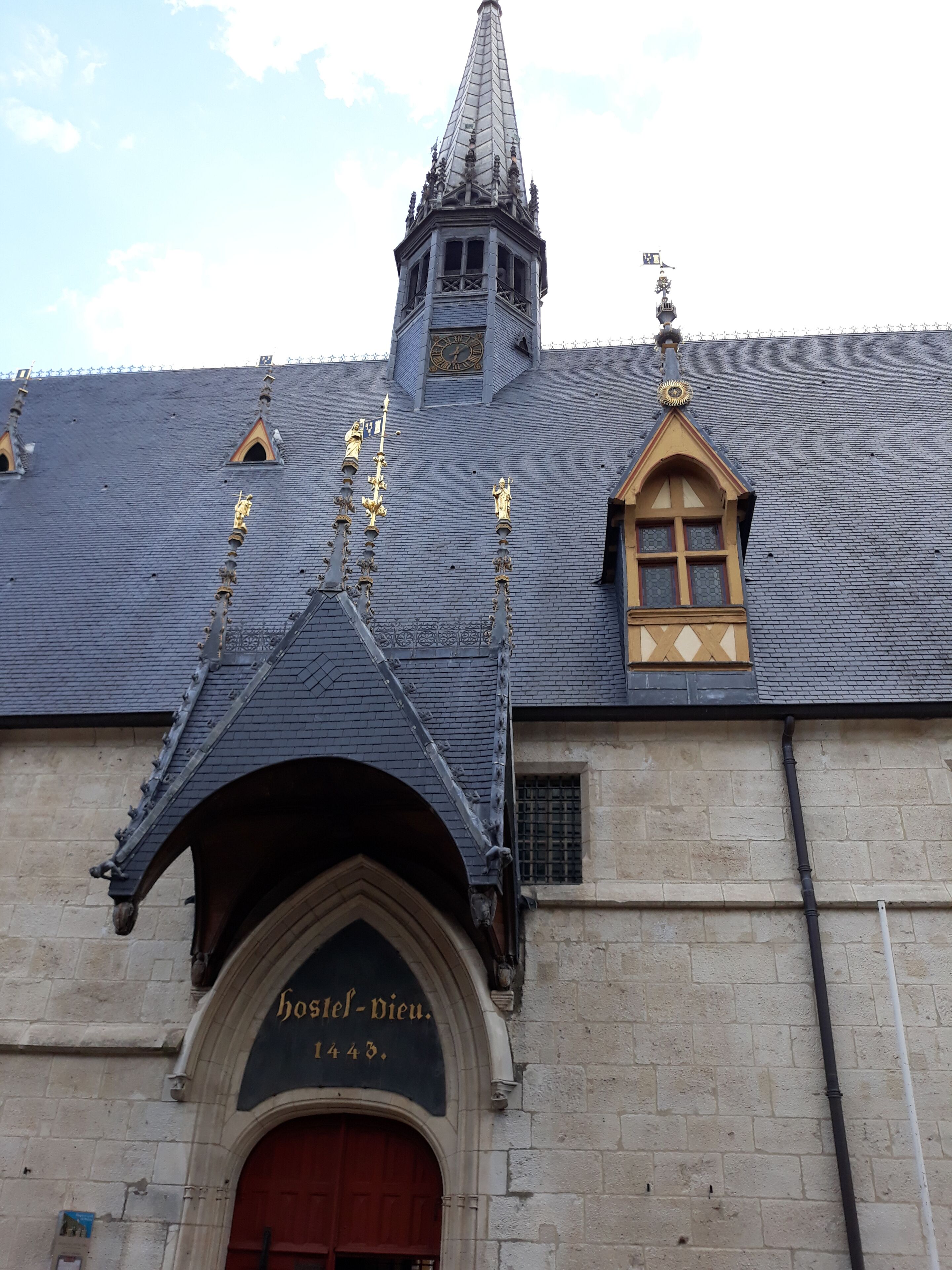 A building with a pointed roof, a clock tower, and a sign reading 'Hostel-Vieu 1443.'
