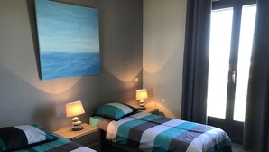 A hotel room with two single beds, a painting on the wall, two bedside lamps, and a view of the outdoors through a window.