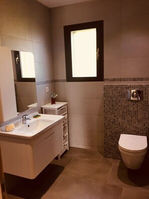 Une salle de bain moderne avec un meuble-lavabo blanc, un grand miroir, une fenêtre et une zone de douche carrelée.