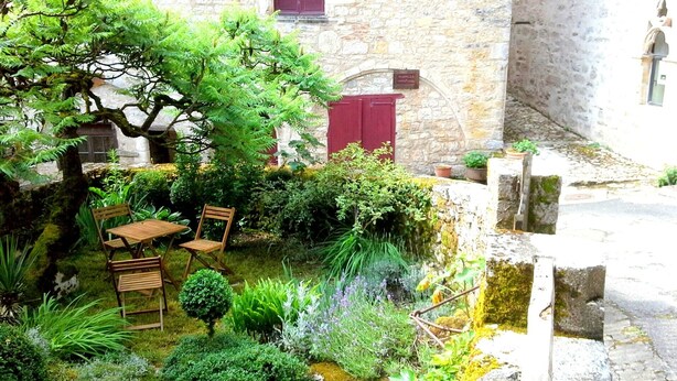 Une cour avec un mur en pierre, une table en bois et des chaises. On y trouve différentes plantes et un petit arbre.