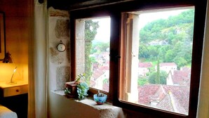 Une pièce avec une fenêtre donnant sur un village, une horloge murale et une petite table avec une lampe.