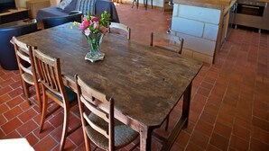 Een ruime woonkamer met een houten eettafel, stoelen en een bank. Er is een keuken met een spoelbak en fornuis.