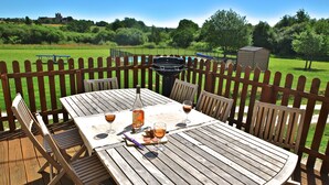 Een houten terras met een ronde tafel en stoelen, een barbecue en een zwembad op de achtergrond.