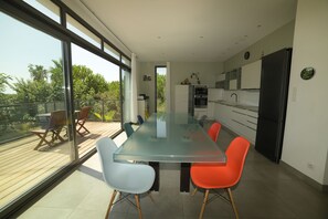 A modern kitchen with a glass dining table and chairs, large windows, and a view of greenery.