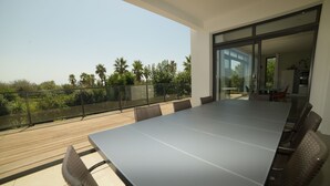 A modern outdoor dining area with a long table and chairs, overlooking a garden with palm trees. - SETE, listed villa 240 m2, 50 meters from the beach, 1st sea line, swimming pool (Sète)