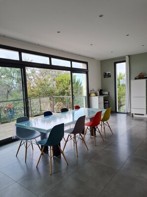 A modern dining area with a glass table and colorful chairs, large windows with a view of greenery, and a kitchen area with cabinets and a sink.