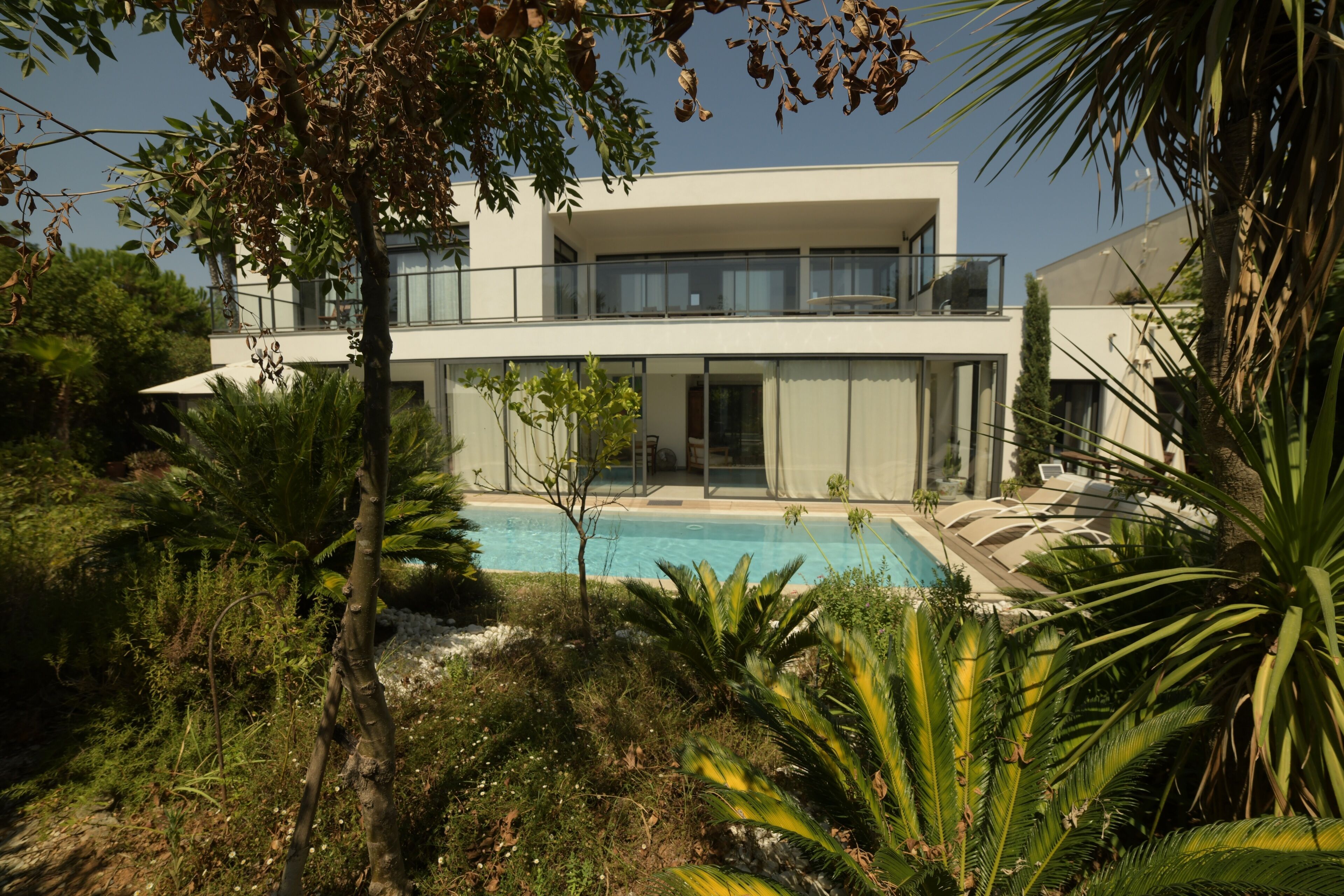 Une maison moderne à deux étages avec une piscine, entourée d’une végétation luxuriante.