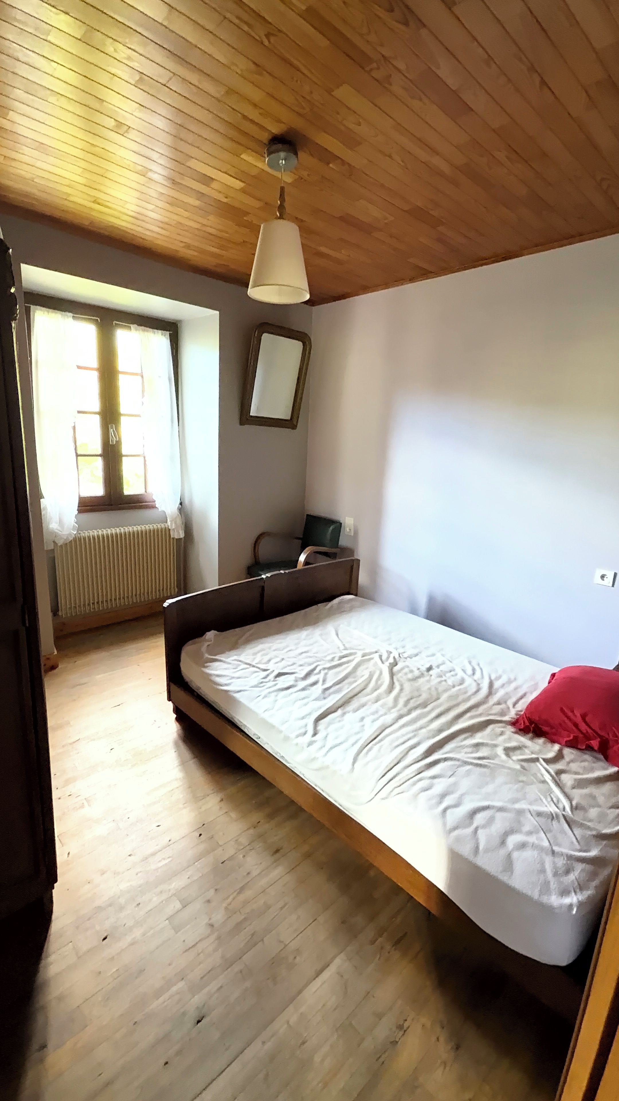 Une pièce avec un plancher en bois, un lit avec un matelas blanc et un oreiller rouge, une chaise, un bureau avec un ordinateur, une fenêtre avec des rideaux et un radiateur.