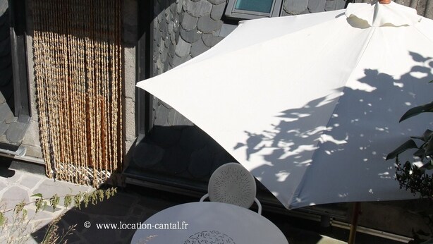 Een terras met een ronde tafel en stoelen, een witte parasol en een stenen muur met een raam.
