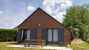 Une maison en bois avec un toit triangulaire, deux grandes fenêtres avec volets, un banc à l’extérieur et un jardinière verte.