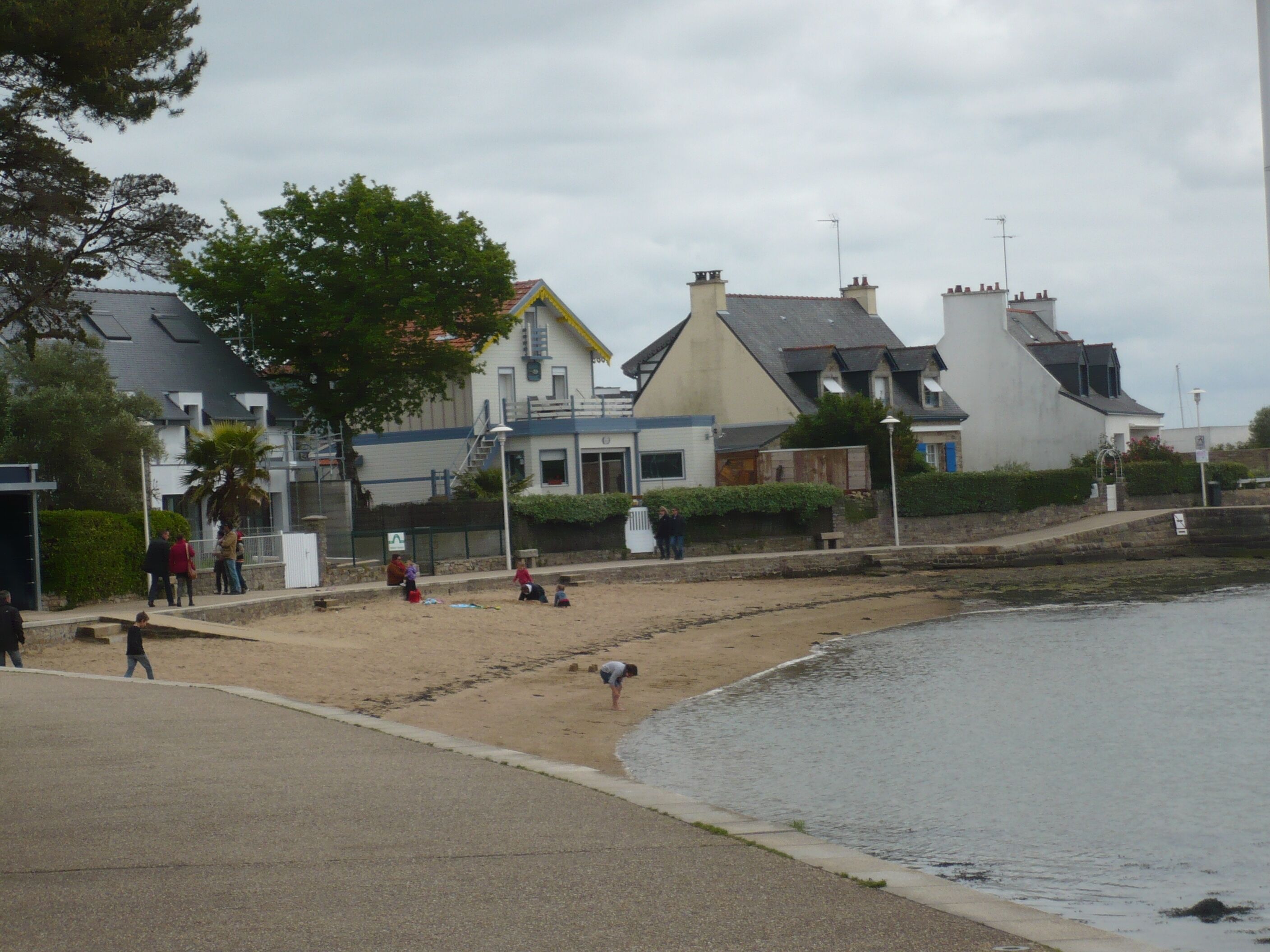 A beach with people, a building with a sloped roof, and a calm body of water.