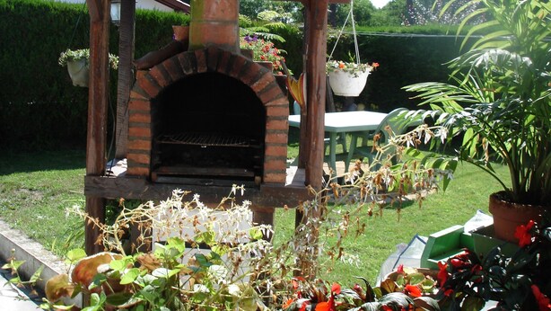 Une structure de four extérieur en briques, surmontée d’un toit en tuiles, entourée de plantes en pot et agrémentée d’une chaise verte.