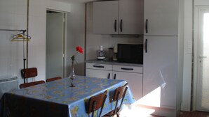 Un coin repas avec une table recouverte d’une nappe à motifs floraux, entourée de chaises. Un vase avec une fleur rouge est posé sur la table. La pièce est dotée de meubles blancs et d’un four à micro-ondes sur le plan de travail.