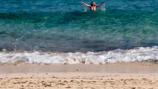 Une scène de plage avec une eau turquoise et limpide, et une personne debout dans l’eau, les bras levés.