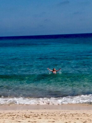 Eine Strandansicht mit klarem türkisblauem Wasser und einer Person, die im Wasser mit erhobenen Armen steht.