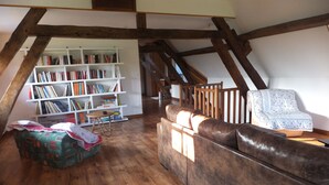 A loft bedroom with a wooden beam ceiling, a built-in bookshelf, a cushioned bench, and a floral-patterned chair.