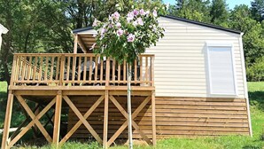 Eine Holzterrasse, an ein weißes Wohnmobil mit Balkon angebaut, umgeben von Gras und Bäumen.