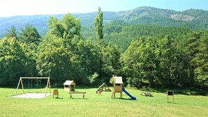 Ein Spielplatz mit Schaukeln, einer Rutsche und einer kleinen Hauskonstruktion in einer Wiese vor einer bewaldeten Berglandschaft.
