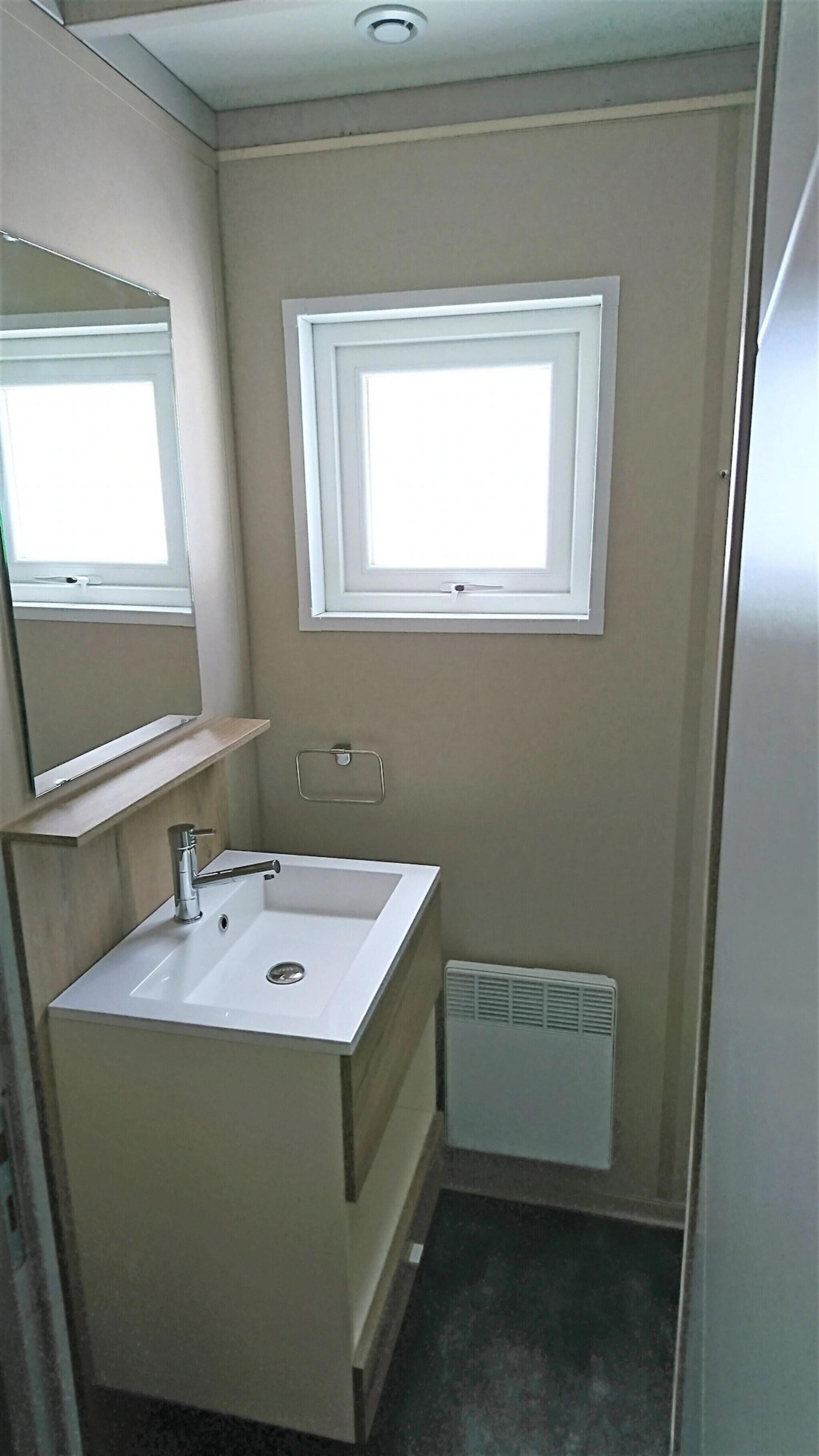 Een compacte badkamer met een wit wastafel, een spiegel, een raam en ventilatie.