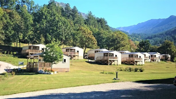 Eine Reihe von Holzhütten mit Terrassen, eingebettet in eine grüne Landschaft mit Bäumen und Bergen im Hintergrund.