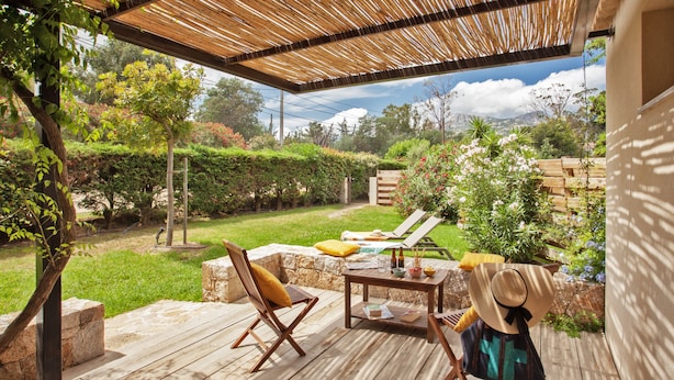 Un patio exterior con suelo de madera, dos sillones, una mesa con un cuenco y vasos, y un sombrero de paja en una de las sillas.