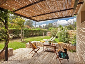Un patio exterior con suelo de madera, dos sillones, una mesa con un cuenco y vasos, y un sombrero de paja en una de las sillas.