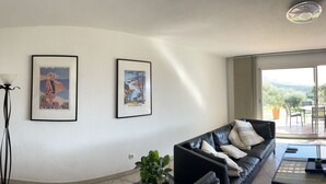 Een moderne woonkamer met een zwarte leren bank, een glazen salontafel en een open haard. Er hangen ingelijste foto's aan de muur en staat een televisie.