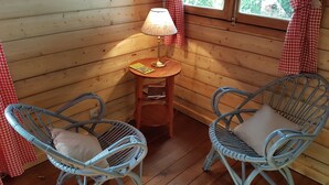 Une pièce chaleureuse aux murs en bois, avec deux fauteuils en osier dotés de coussins, une petite table en bois surmontée d’une lampe, et une fenêtre ornée de rideaux rouges.