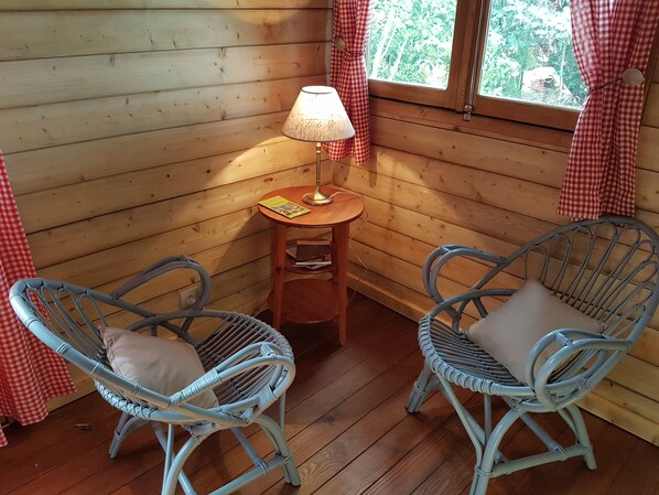 A cozy room with wooden walls, two wicker chairs with cushions, a small wooden table with a lamp, and a window with red curtains. - Tree house in a litchi (sainte suzanne)