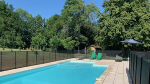 Ein rechteckiger Pool mit Fliesenveranda, umgeben von einem schwarzen Zaun. Im Hintergrund befinden sich zwei grüne Liegestühle und eine kleine Laube.
