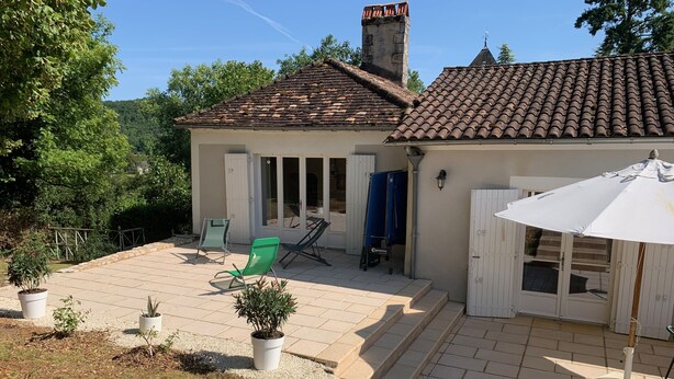 Ein Haus mit Ziegeldach, einer überdachten Terrasse mit weißem Sonnenschirm und einem Garten mit Topfpflanzen.
