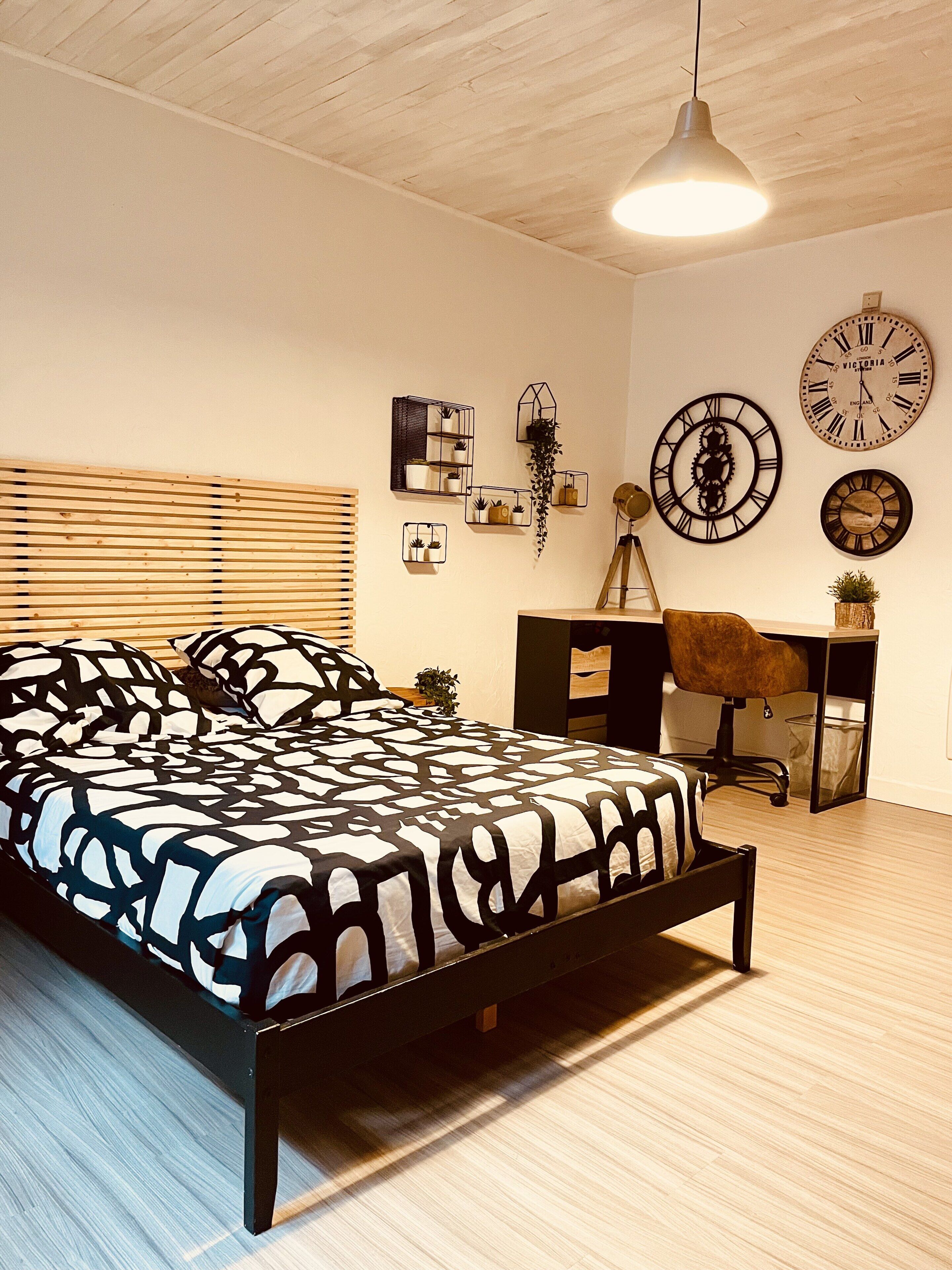 A bedroom with a bed, a desk, and two clocks on the wall.