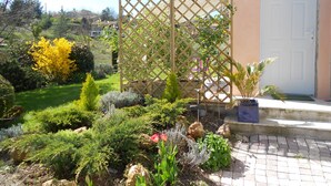 Um jardim com cerca de madeira treliçada, diversas plantas e uma casa com porta branca.
