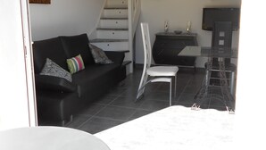 Uma sala de estar com um sofá preto, uma poltrona branca, uma mesa pequena e uma escada que leva a um andar superior.
