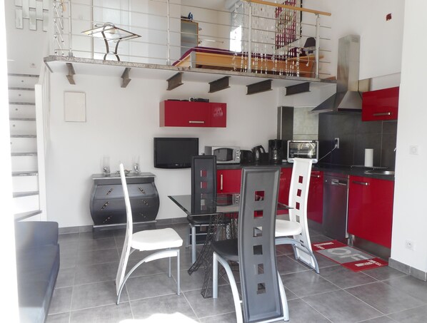 Une cuisine compacte avec des meubles rouges, un plan de travail noir et un coin repas comprenant une table et des chaises en verre.