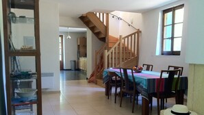 Un comedor con una mesa de madera, sillas y un mantel colorido. Una escalera conduce a otra habitación.