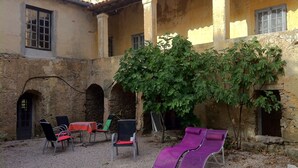 Un área exterior con sillas y mesas, una pérgola con plantas trepadoras y un edificio de piedra con arcos.