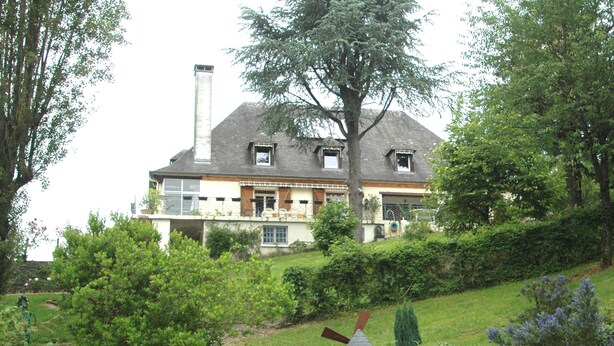 A large house with a chimney, surrounded by lush greenery and a windmill. - The Hydrangeas suite - quality services (in the countryside in Lourdes) (Lourdes)