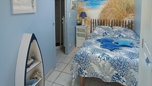 A bedroom with a beach mural, a bed with a blue and white patterned bedspread, a small shelf unit, and a ceiling fan.