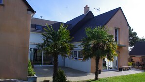 A two-story house with a sloped roof, a balcony, and a small garden with palm trees. - Seaside guest room 1 (Portbail)