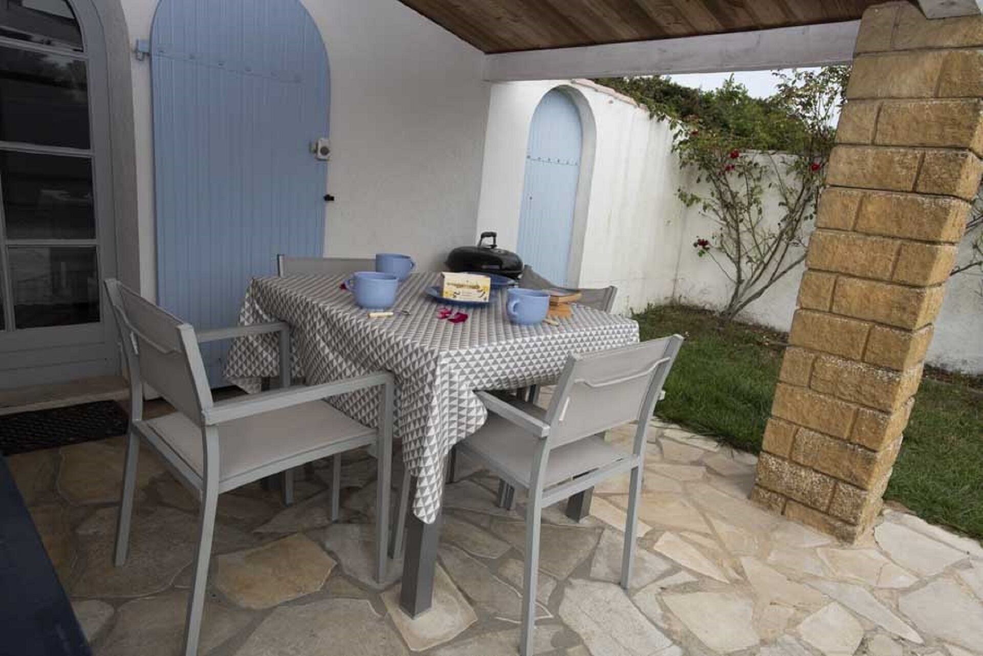 En udendørs terrasse med et bord dækket til to, omgivet af stenbelægning og en hvid væg med en blå dør.