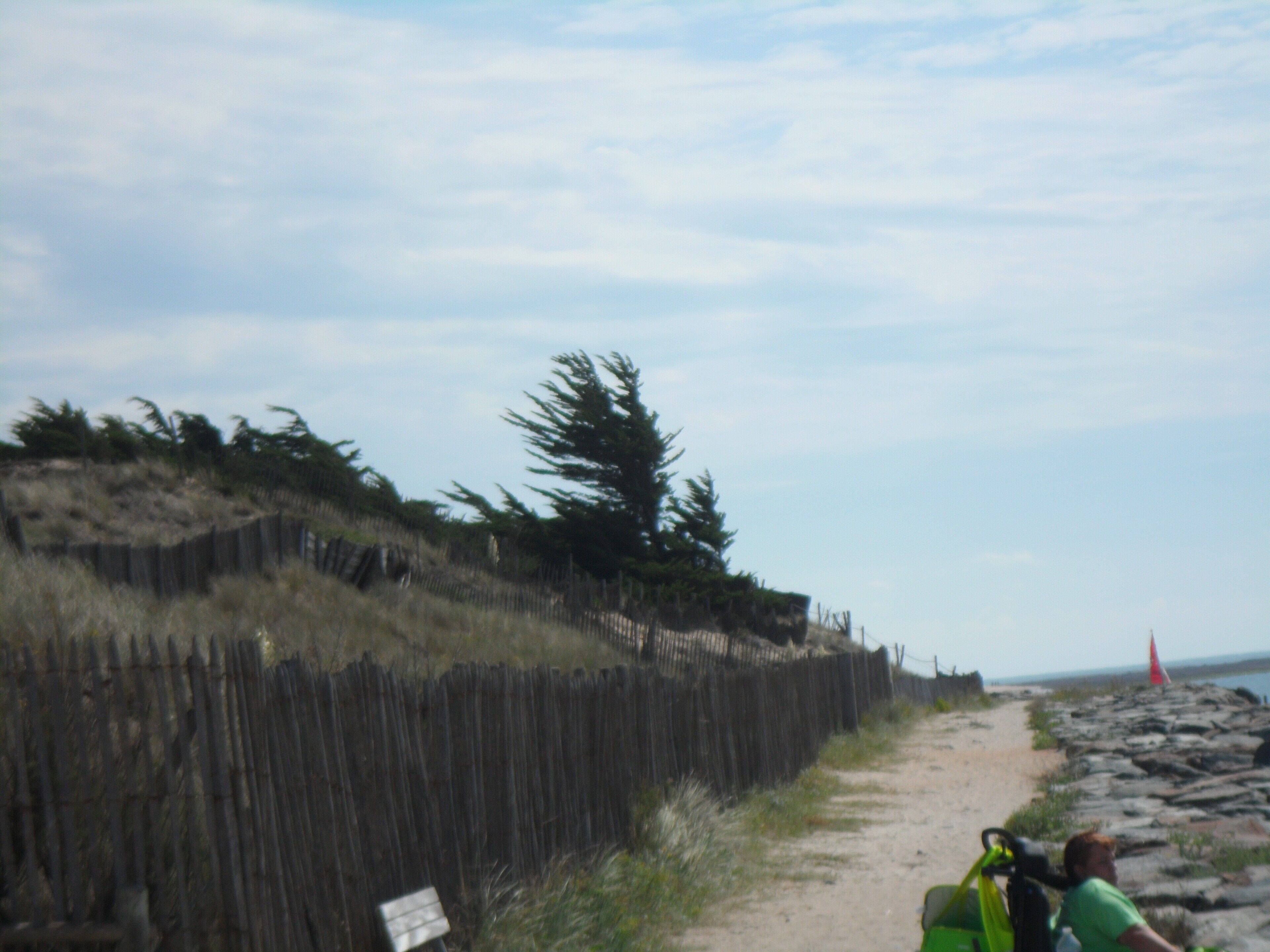 A dirt path beside a wooden fence with a sign, leading to a beach with a red flag.