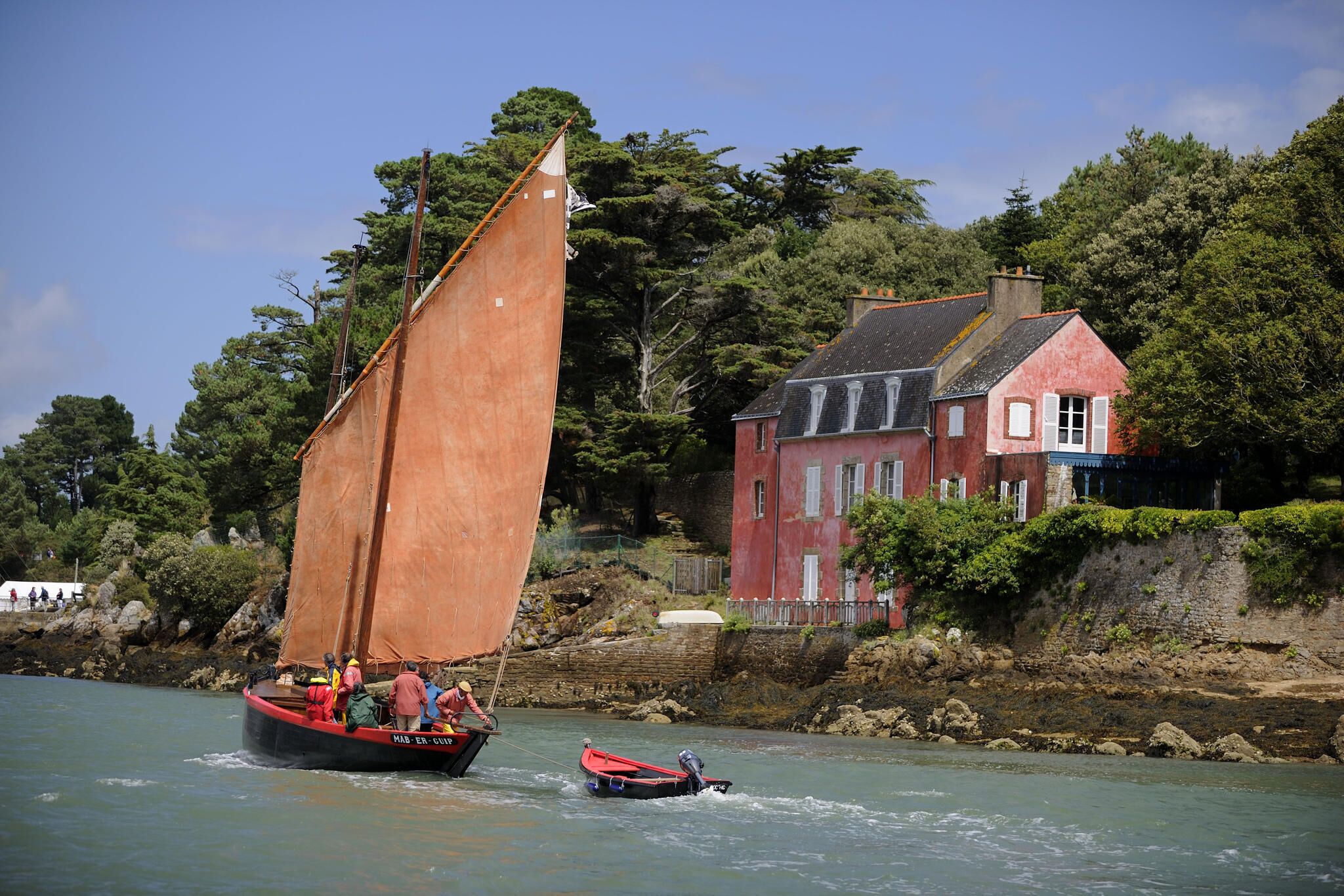 Un voilier transportant des passagers, un petit bateau à moteur et une maison rouge au bord de l’eau.