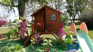 A wooden playhouse with a small balcony, surrounded by green foliage and pink flowers. - Small country house in the heart of the vineyards DU (Fronton)