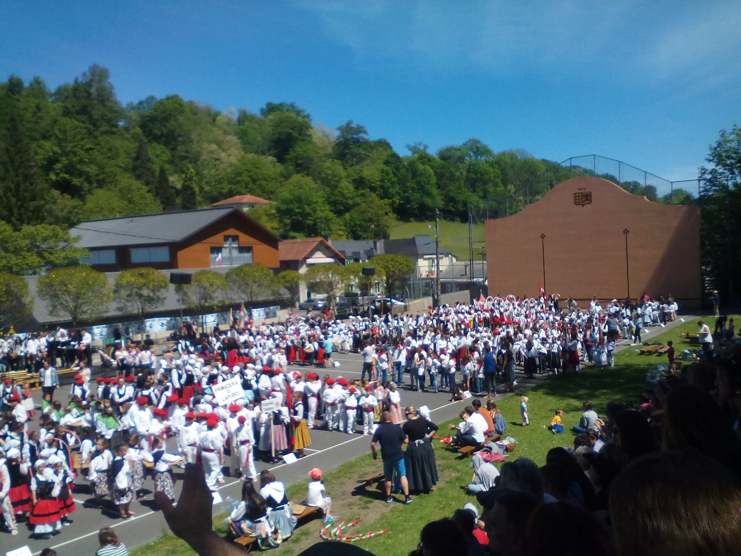 Une grande fête en plein air où les participants sont vêtus de costumes traditionnels, un bâtiment à la façade marron et un ciel bleu dégagé.