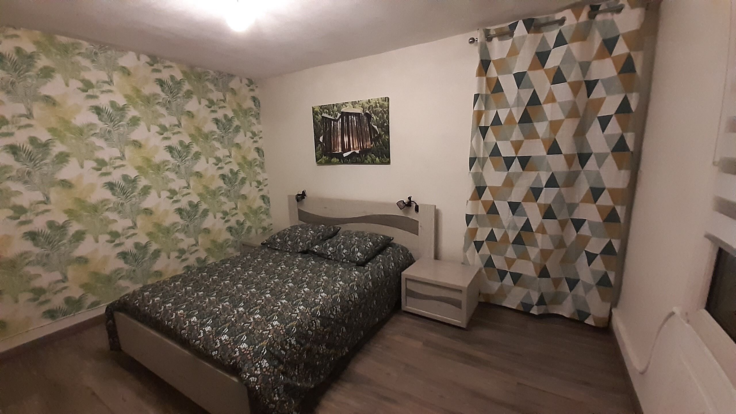 Une chambre à coucher comprenant un lit, une table de chevet, un tableau accroché au mur et un rideau de douche à motifs géométriques.
