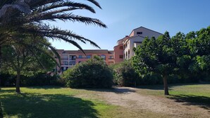 A courtyard with a palm tree, a bush with pink flowers, and a building with multiple balconies. - Studio cabin 4 pers, in quiet residence with swimming pool, 50 m from the beach (Saint-Cyprien)