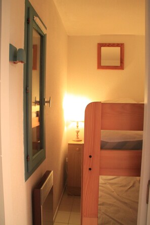 Ein schmales Zimmer mit einem Holzbett, einem kleinen Nachttisch mit Lampe, einem Spiegel an der Wand und einem Heizkörper.