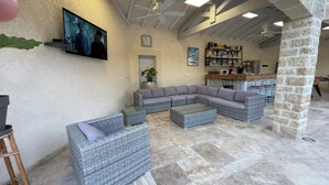Amplia sala de estar con un sofá modular grande, dos sillones y una mesa de centro. Un televisor de pantalla plana está montado en la pared, y hay una zona de bar con estantes llenos de vasos y botellas.